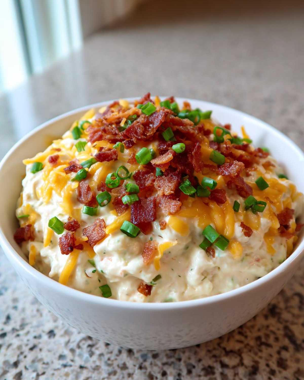 A creamy bowl of Million Dollar Dip topped with shredded cheddar cheese, bacon bits, and green onions.