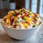 A creamy bowl of Million Dollar Dip topped generously with shredded cheddar cheese, bacon bits, and sliced green onions.