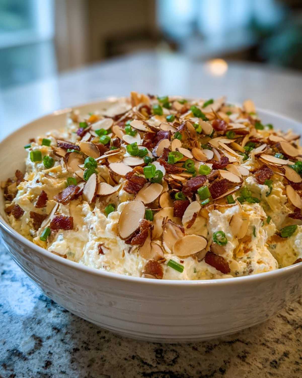 A large white bowl filled with creamy Neiman Marcus dip, topped generously with sliced almonds, bacon bits, and chopped green onions.
