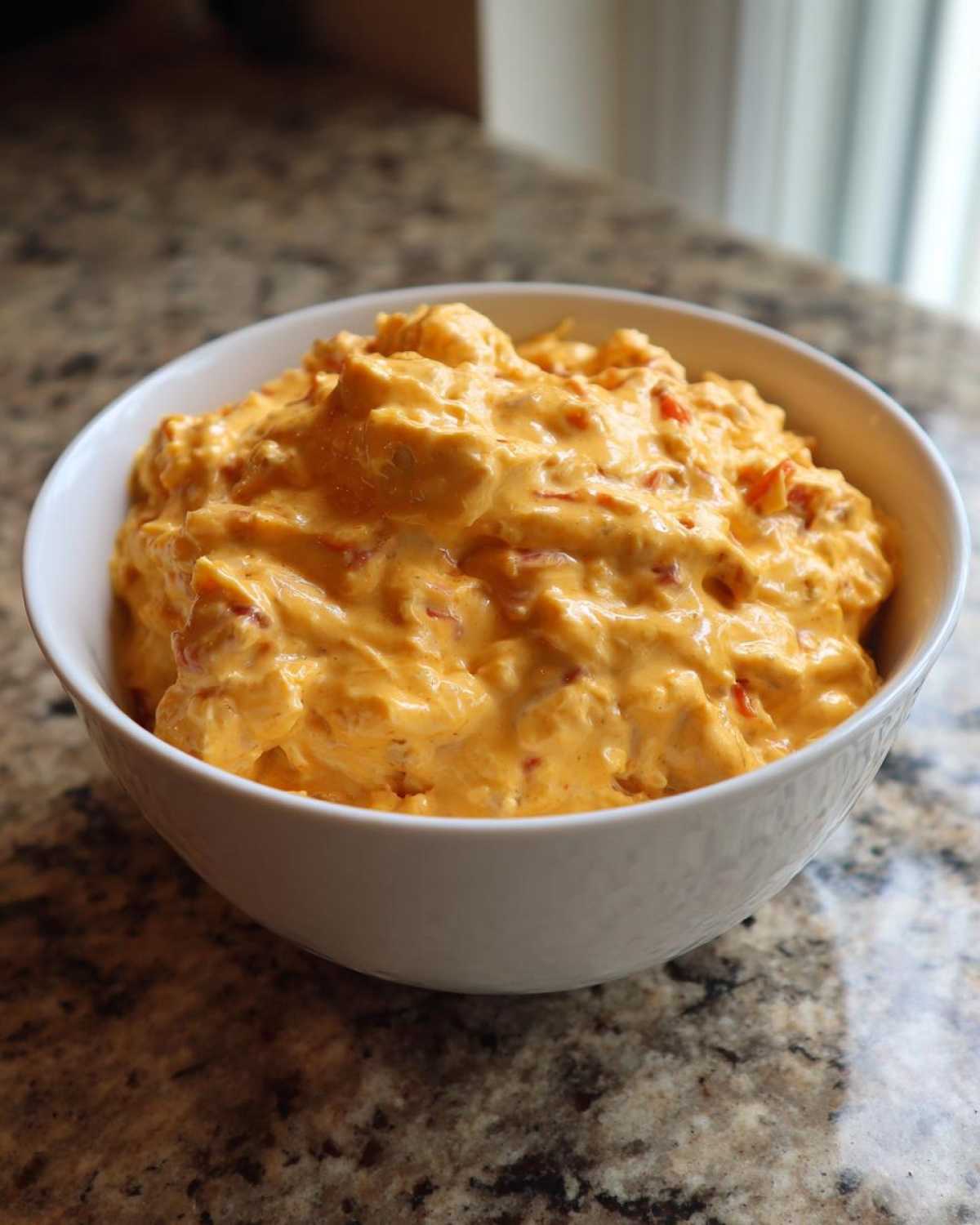 A white bowl filled with creamy, orange Rotel dip featuring visible pieces of diced red peppers.
