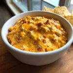 A close-up of hot, creamy rotel dip with chunks of meat and peppers served in a white bowl with tortilla chips.