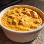 Close-up of hot, creamy orange Rotel dip served in a small white bowl on a granite countertop.