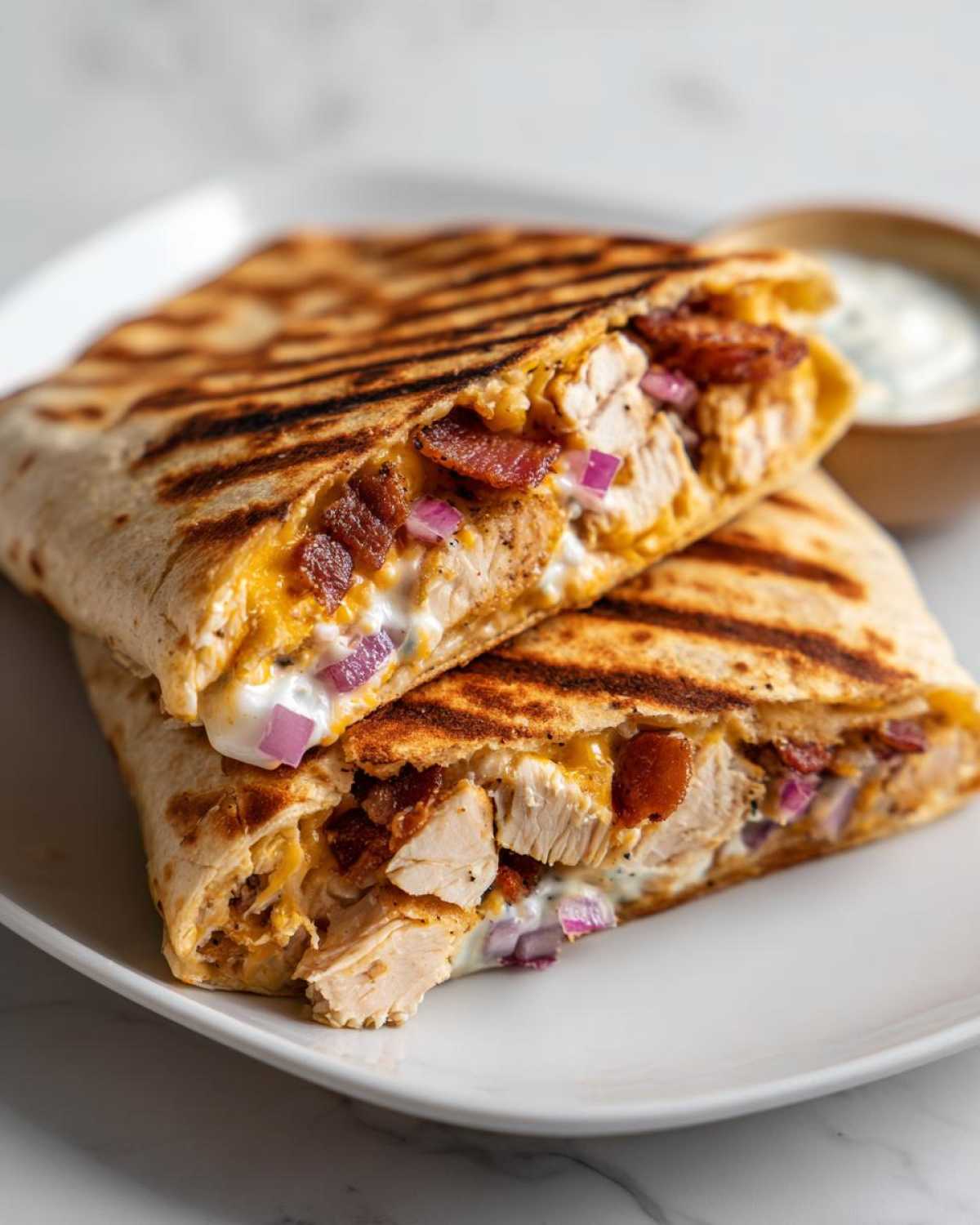 A grilled chicken bacon ranch wrap cut in half, showing chunks of chicken, bacon, cheese, and red onion inside.