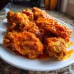 Close-up of several crispy chicken wings heavily coated in bright orange buffalo sauce on a white plate.