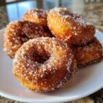 Close-up of several golden brown, crispy buñuelos heavily coated in sparkling cinnamon sugar on a white plate.