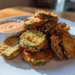 A pile of golden brown, crispy fried pickles served on a white plate next to a small bowl of creamy dipping sauce.