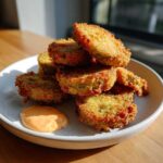 A stack of golden brown, crispy Fried Pickles served on a light speckled plate with a side of orange dipping sauce.