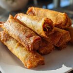 A stack of golden brown, crispy fried egg roll appetizers piled on a white plate.