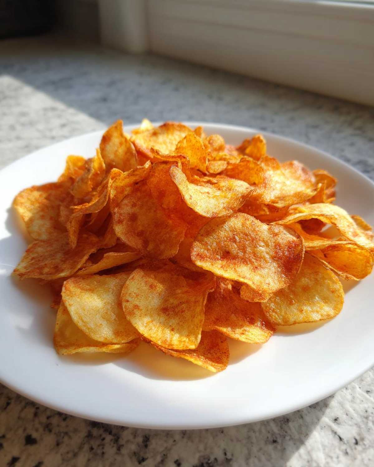A pile of golden, crispy potato chips seasoned with paprika sits on a white plate near a window.