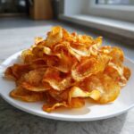 A white plate piled high with golden, crispy homemade potato chips seasoned with paprika.
