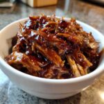 Close-up of shredded crockpot teriyaki chicken coated in glossy, dark teriyaki sauce, served in a white bowl.
