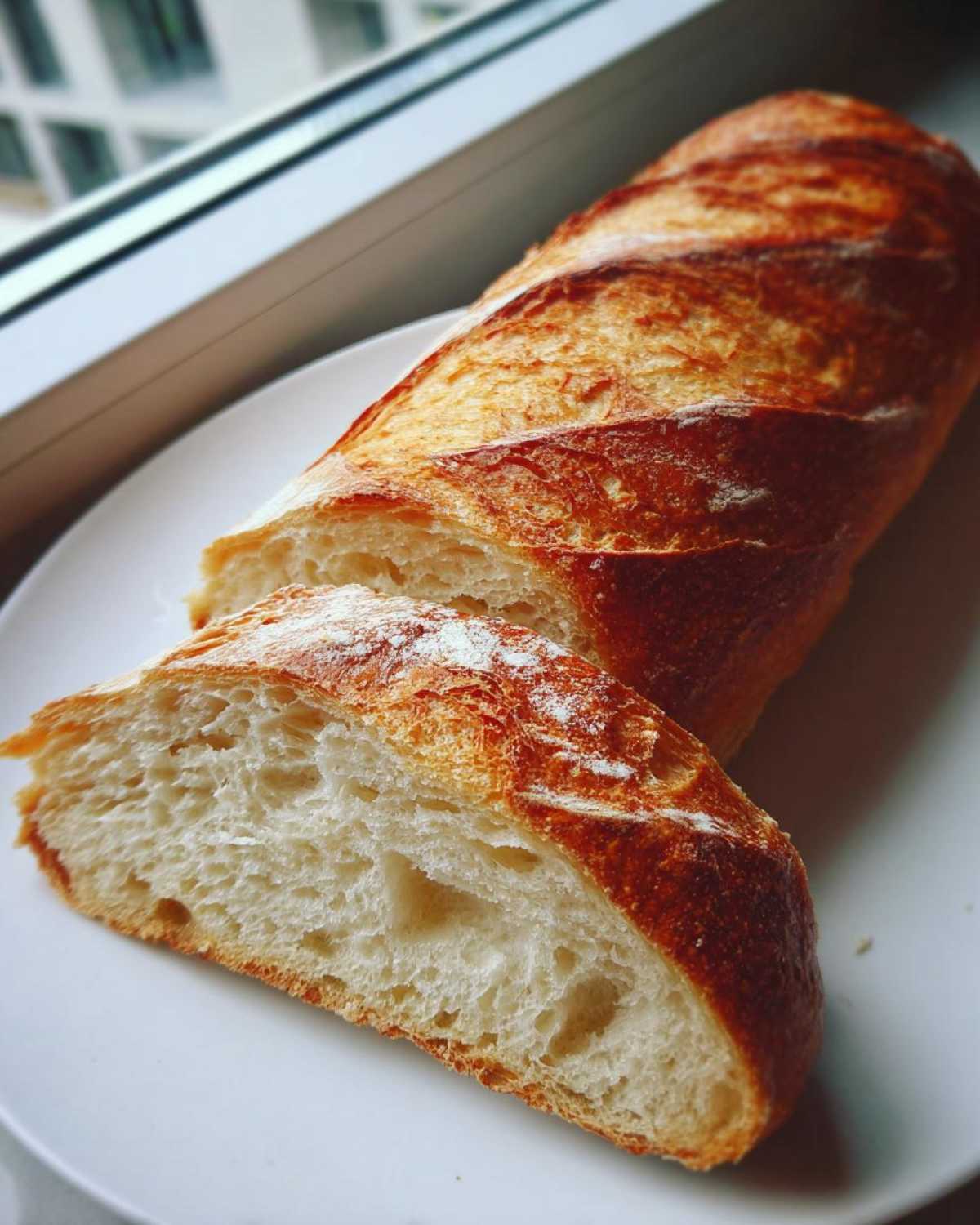 A golden-brown, crusty loaf of homemade french bread, partially sliced on a white plate.