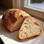 A loaf of homemade french bread sliced open, showing the airy interior and crust, set against a bright window.