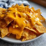 A close-up shot of crispy tortilla chips generously smothered in bright orange, smooth nacho cheese sauce.