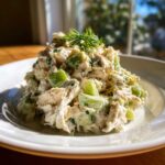 A mound of creamy dill pickle chicken salad featuring chunks of chicken and bright green pickles, topped with fresh dill.