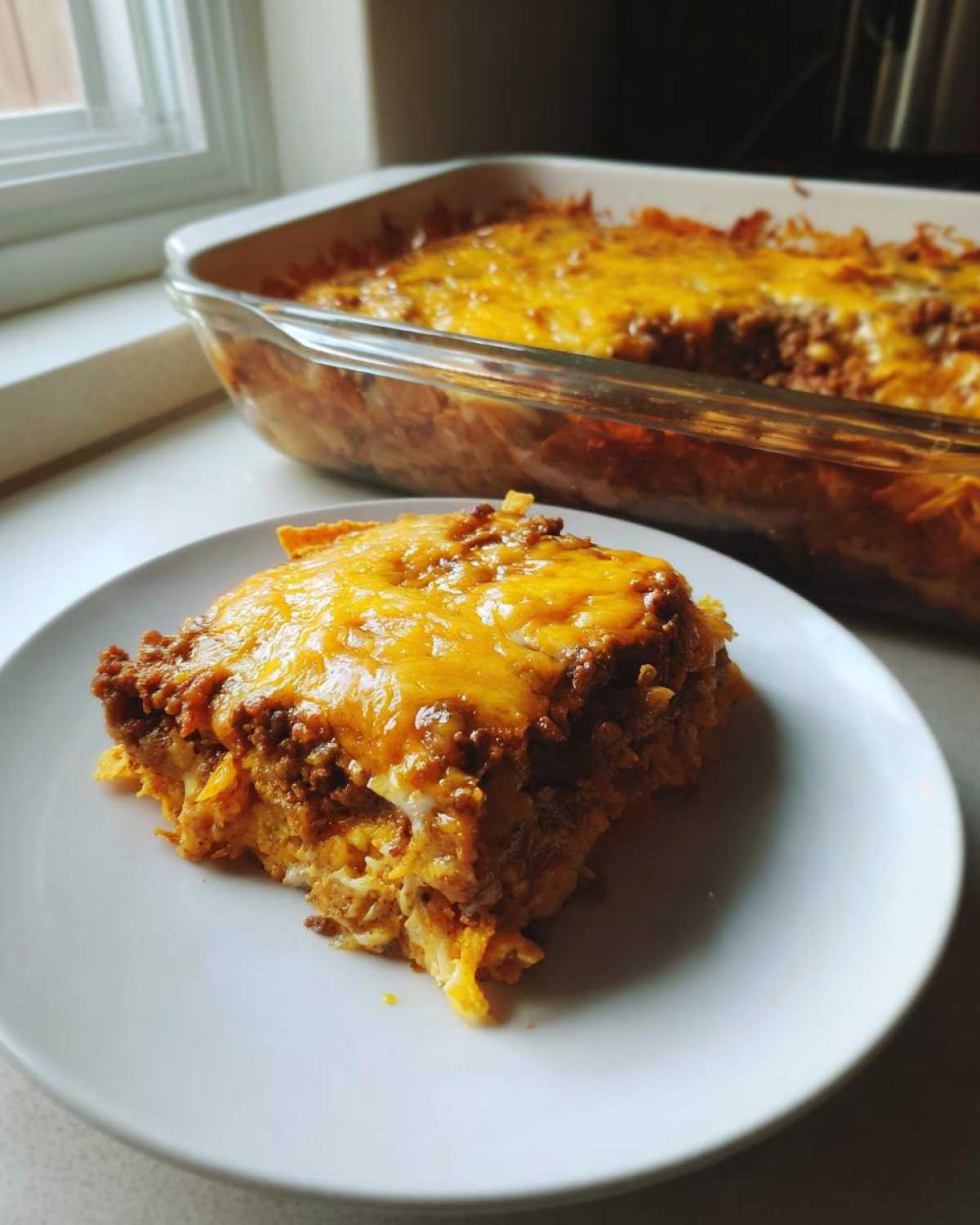 A serving of Dorito Casserole topped with melted cheddar cheese, with the baking dish visible in the background.