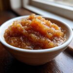 Close-up of chunky, spiced homemade applesauce served in a small white bowl near a window.
