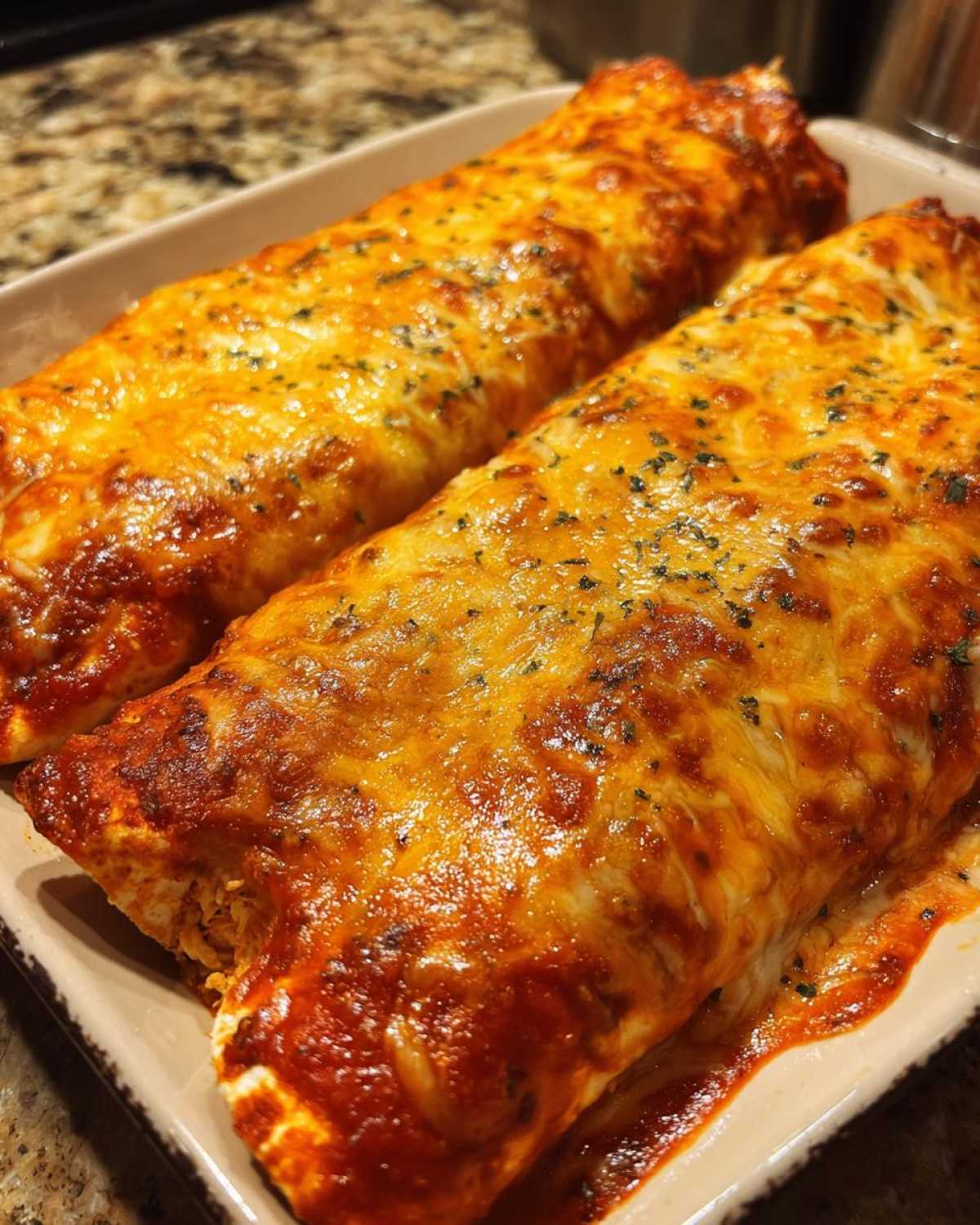 Two baked easy chicken enchiladas covered in bubbly, melted cheese and red sauce, sprinkled with herbs.