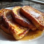 Three golden slices of easy french toast dusted with powdered sugar and drizzled with syrup on a white plate.