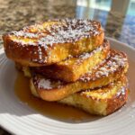 A stack of four slices of golden brown easy french toast dusted heavily with powdered sugar and drizzled with syrup.