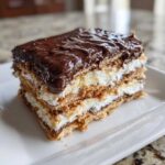 A close-up of a layered slice of eclair cake with graham crackers, creamy filling, and thick chocolate frosting.