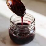 A spoonful of thick, dark elderberry syrup is dripping into a small glass jar, illustrating the elderberry syrup recipe.