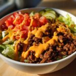 A close-up of a vibrant cheeseburger bowl featuring seasoned ground beef, lettuce, diced tomatoes, pickles, and a generous drizzle of orange sauce.