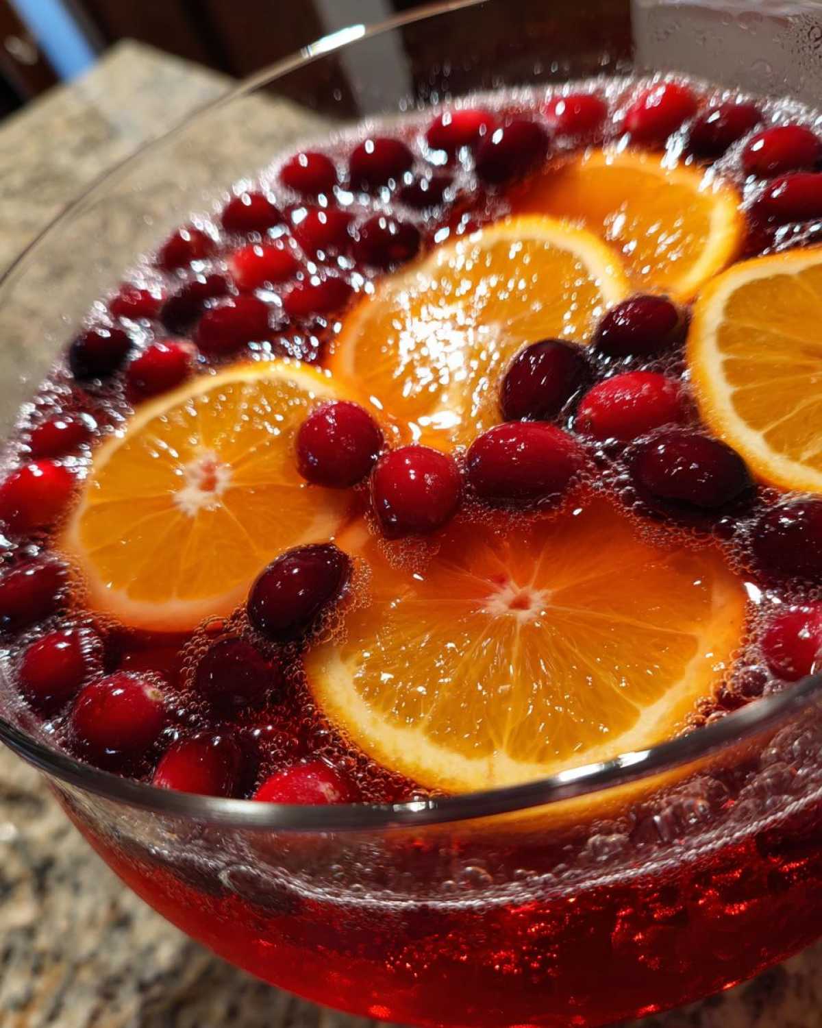 Close-up of a sparkling red champagne punch garnished with bright orange slices and whole cranberries in a glass bowl.