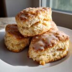 Close-up of three flaky, buttery scones stacked on a white plate, drizzled with a sweet white glaze.
