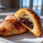 A flaky, golden-brown chocolate croissant cut in half, revealing a rich, melted chocolate filling inside.