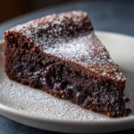A close-up of a rich slice of flourless chocolate torte, dusted heavily with powdered sugar.