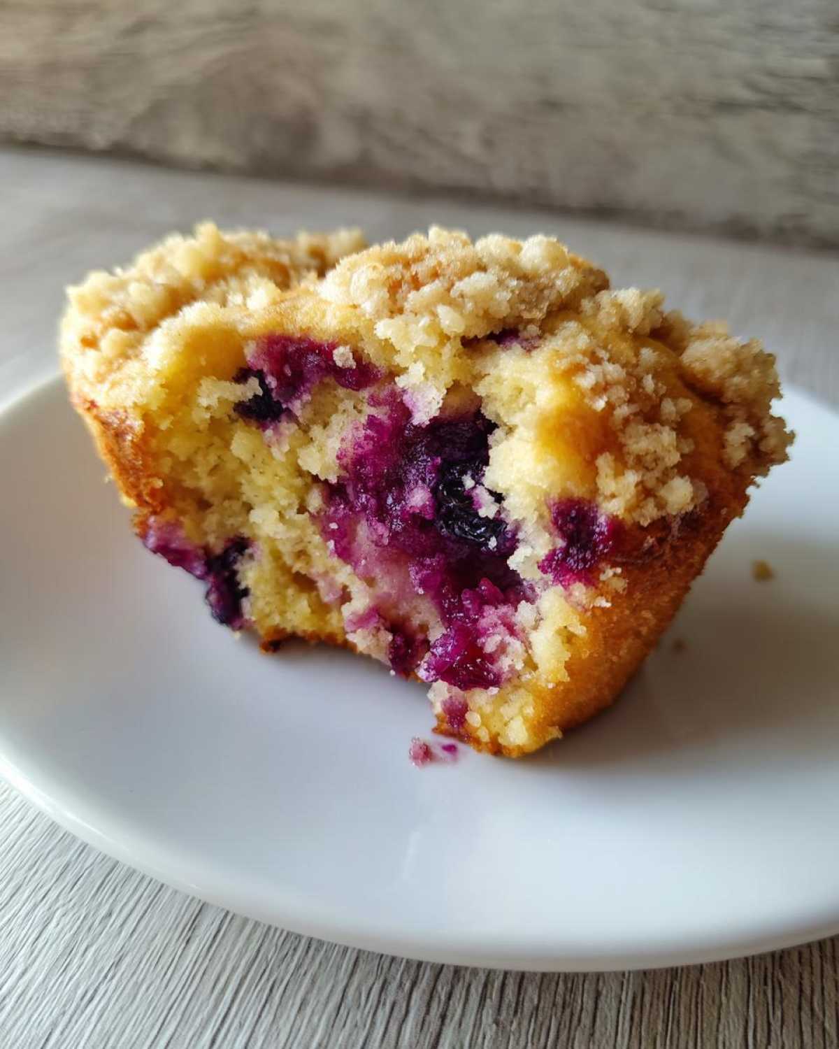 A close-up view of a fluffy blueberry muffin cut in half, showing juicy berries and a crumb topping, resting on a white plate.