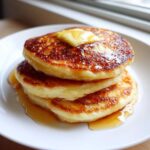 A stack of three golden-brown greek yogurt pancakes topped with melting butter and drizzled with syrup.