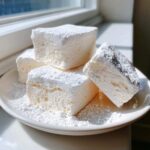 Four thick, square pieces of fluffy homemade marshmallows dusted heavily with powdered sugar on a white plate.