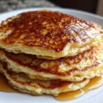 A close-up of a tall stack of fluffy oatmeal pancakes drizzled generously with golden syrup.