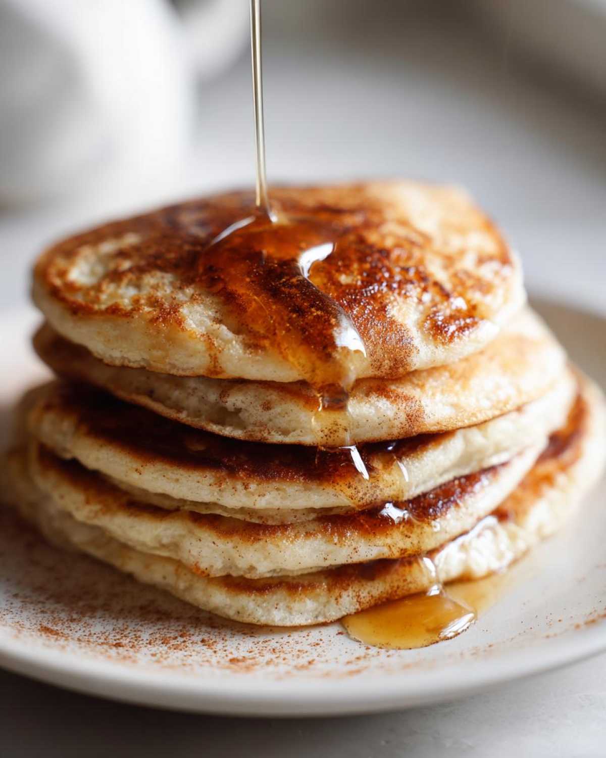 A tall stack of fluffy oatmeal pancakes being drizzled with maple syrup, sprinkled with cinnamon.