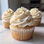A close-up of one perfectly frosted vanilla cupcakes with two others blurred in the background.