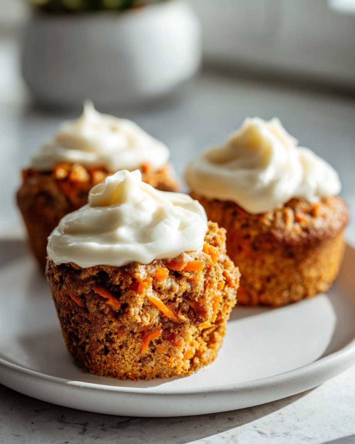 Three moist carrot cake muffins topped with thick cream cheese frosting, sitting on a white plate.