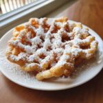 A golden brown, freshly fried funnel cake recipe dusted heavily with powdered sugar on a white plate.