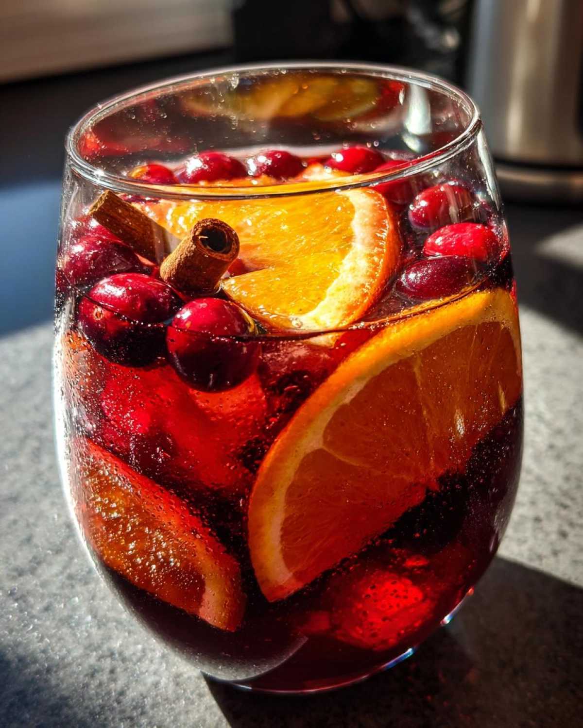 Close-up of a glass filled with vibrant red Christmas sangria, garnished with orange slices, whole cranberries, and a cinnamon stick.