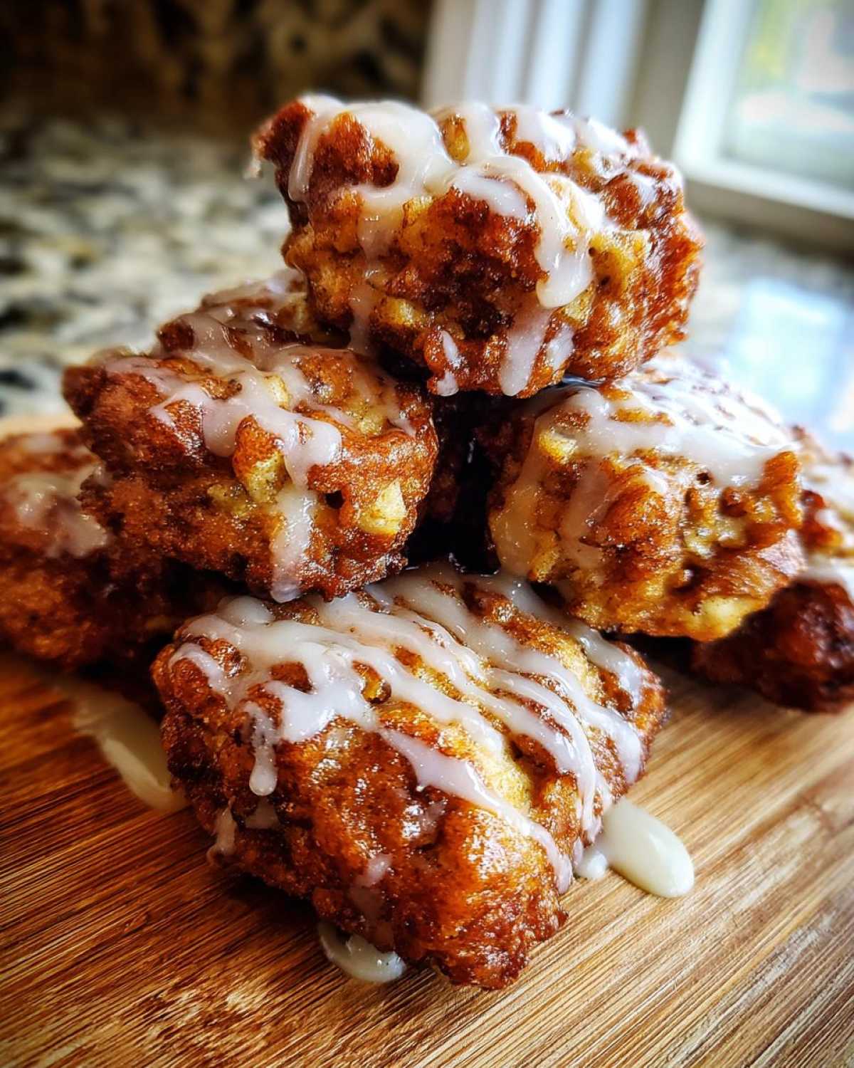 A stack of golden brown, glazed apple fritter bites drizzled with white icing, sitting on a wooden cutting board.