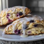 Two freshly baked blueberry scones, drizzled with a thick white glaze, resting on a white plate.