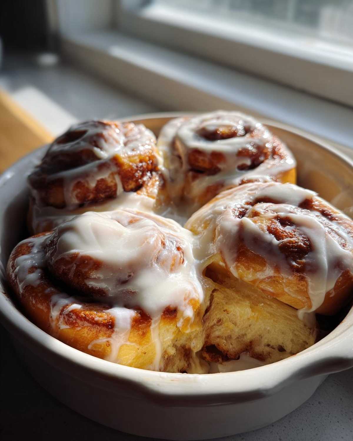 Three freshly baked cinnamon roll swirls topped with thick white icing, sitting closely together in a white baking dish.