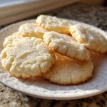 A plate piled high with soft, round vegan sugar cookies topped with a light glaze, catching warm sunlight.