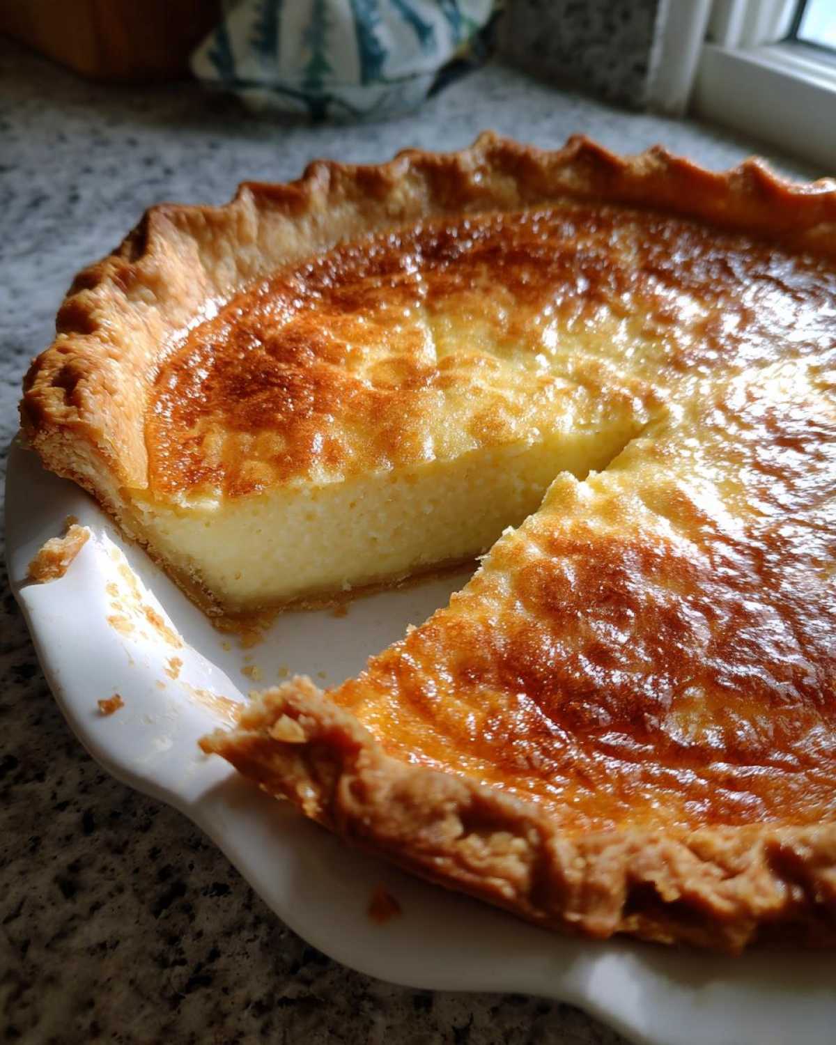 A freshly baked buttermilk pie with a deeply browned, glossy top, showing a creamy yellow interior where one slice has been removed.