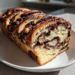 A freshly baked, golden chocolate babka loaf resting on a white plate, showing rich chocolate swirls inside.