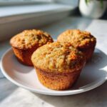 Three freshly baked morning glory muffins with visible shredded carrots sitting on a white plate.