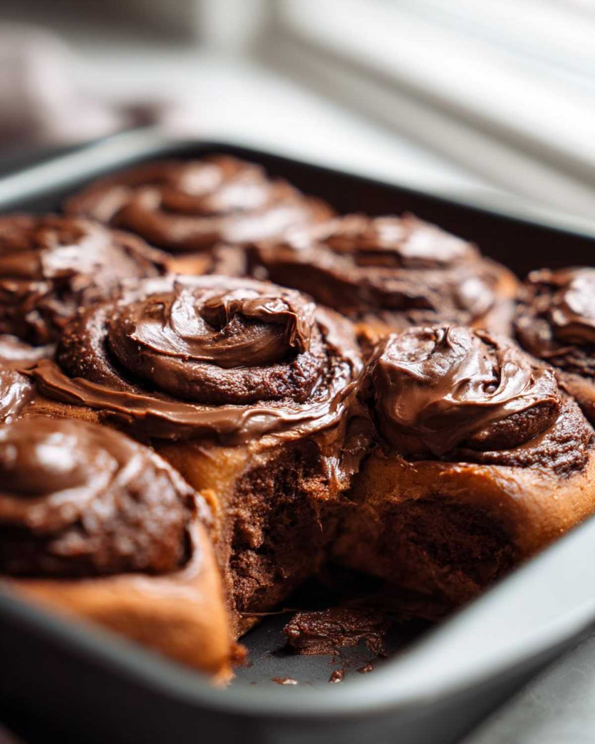 Close-up of gooey chocolate cinnamon rolls topped with thick chocolate frosting, one roll partially pulled apart.