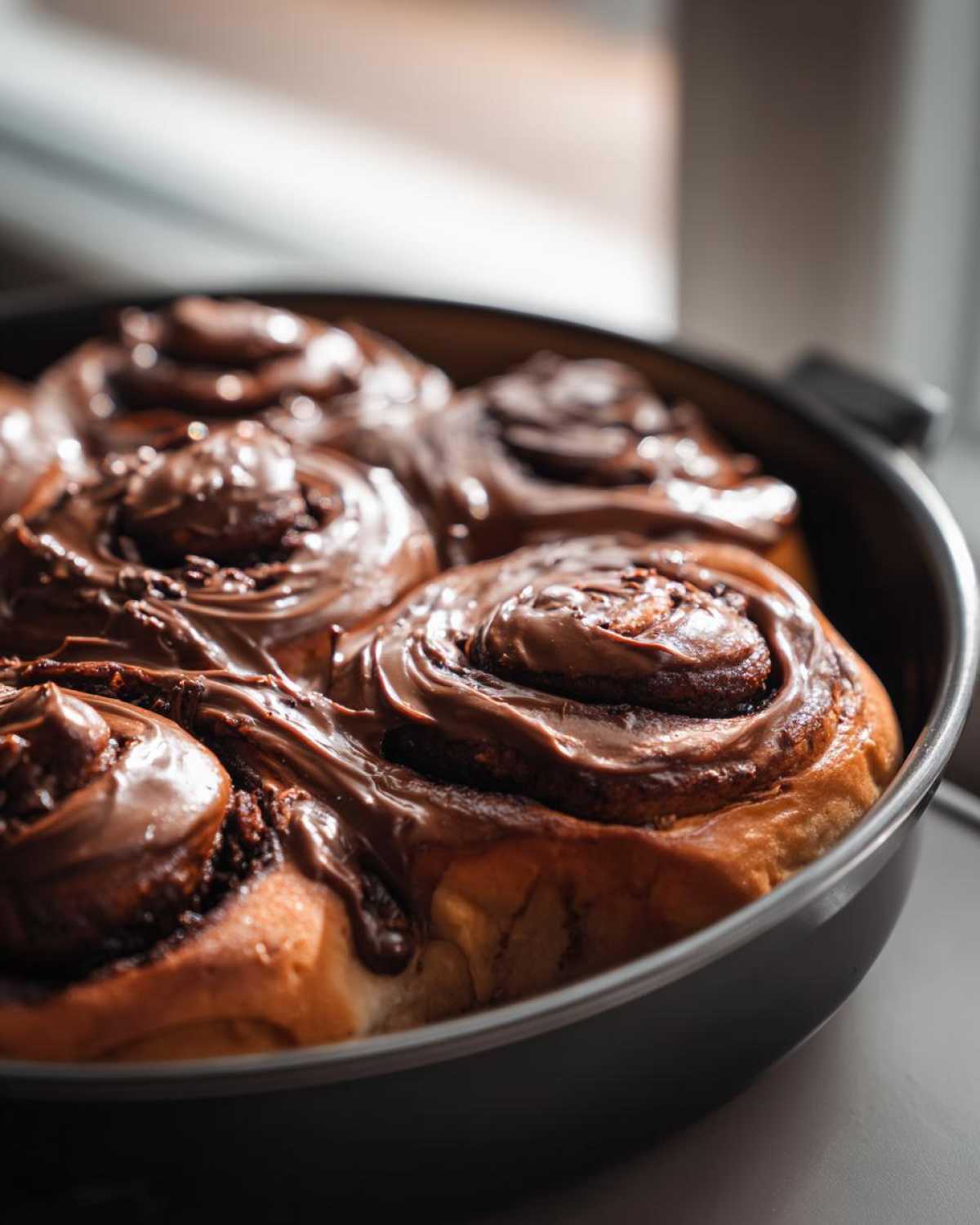 Close-up of several gooey chocolate cinnamon rolls covered in thick chocolate frosting in a baking dish.
