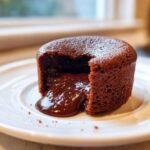 Close-up of a rich chocolate lava cake with a piece removed, showing the molten chocolate center flowing out onto a white plate.